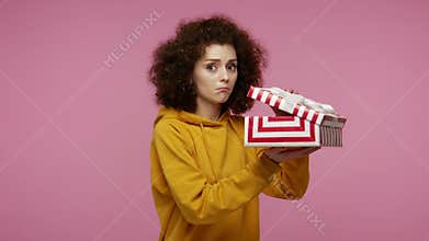 Upset girl afro hairstyle in hoodie opening gift box, unwrapping birthday present and expressing disappointment