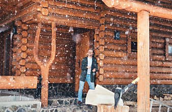 Man staying in the country house porch and enjoying snowfall. Wooden stump with axe on snowy yard unfocused with heavy snowflakes