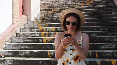 Young woman with braces in straw hat and black sunglasses standing on steps with phone in hands and smiling