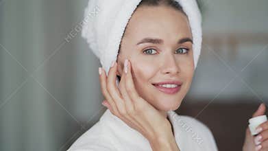 Woman with towel on head applies moisturizer cream on her face, close up