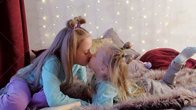 Children girls sisters kissing playing together on bed in bedroom with garland.