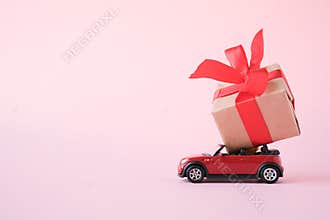 Red toy car delivering gift box with red ribbon on pink background. Place for text. Sales, Birthday, christmas time. St