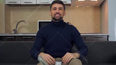 Happy young man in dark blue pullover and gray pants is sitting on the couch and throwing a lot of money in the air