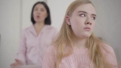 Close-up of young irritated caucasian girl listening to adult brunette woman shouting at the background. Annoyed mother