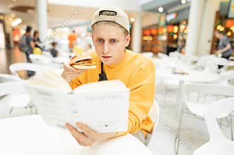 Portrait of a concentrated young man in casual clothing and caps, read a book and eat a sandwich. Boy with a burger in his hand