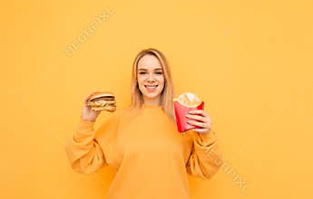 Portrait of a happy girl blond girl standing on a yellow background in an orange casual clothing with burger and french fries in