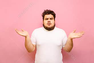 Puzzled young overweight man stands on a pink background and spreads his arms out to the sides. Confused fat guy does not know,