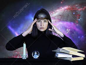 A girl in black clothes sitting at a table with books, a sphere and a burning candle against background of starry sky