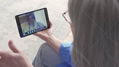 Over shoulder view of old woman video calling virtual doctor on tablet at home.