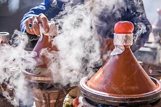 A hand of man cooking traditional Tajine in steam vapor on an outdoor barbecue at town square on the street in Morroco