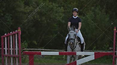 Slow motion, close up: young horsegirl horseback riding strong brown horse jumping the fence in sunny outdoors sandy