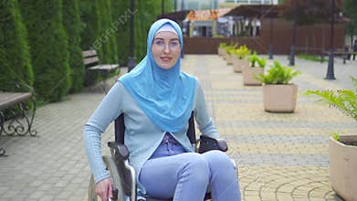 Portrait smiling young muslim woman disabled in traditional scarf sitting in a wheelchair in the park