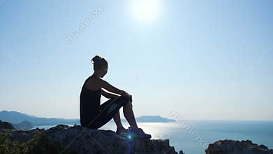 Young inspired woman sitting on the top of a mountain above the sea against beautiful blue sky. Silhouette of a happy