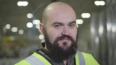 Camera moving up, portrait of bearded baldheaded Caucasian man smiling at camera. Happy worker in green vest standing at