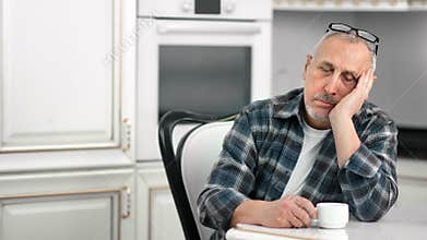 Tired sluggish aging male dozing during drinking tea at white kitchen medium shot