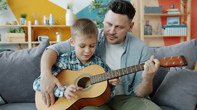 Adorable boy playing the guitar under guidance of caring father learning at home