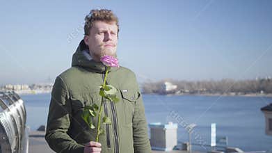 Portrait of young Caucasian redhead man with mustache and beard waiting for sweetheart holding rose. Boyfriend standing