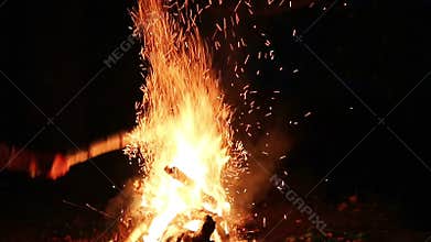 Slow motion - Bonfire by the river in the forest at night. A man bangs a stick on a fire, sheaves of sparks