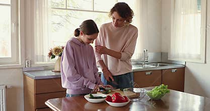 Teen daughter helping young mom cutting fresh salad in kitchen