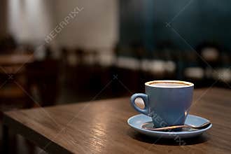 A blue cup of coffee resting on a wooden cafe table against a backdrop of ambient lighting.