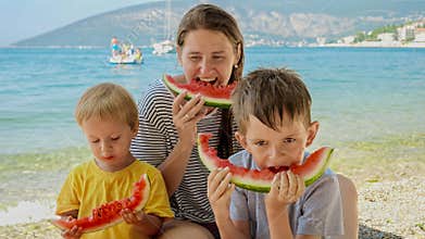 Happy mother and her two boys share laughter and sweet watermelon on a beachside picnic. Family vacation, the happiness