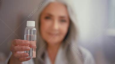 Old gray-haired elderly lady Caucasian woman in bathrobe shows advertises bottle with tonic serum oil with vitamins