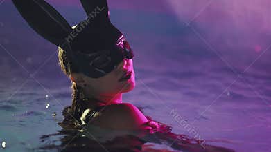 Sexy woman in bunny latex mask swimming in pool at night. Attractive chick in black swimsuit enjoying party time. Rich