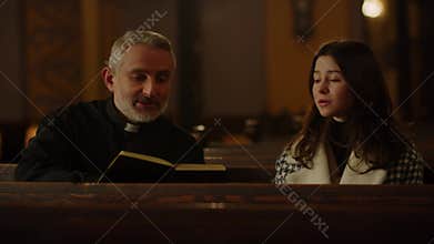 A Catholic priest sitting with a bible in his hands listens to the confession of a brunette woman. A chaplain reads a