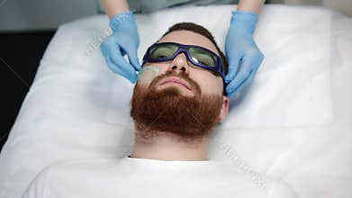 Beautician has applied gel to his face and is putting on protective goggles.