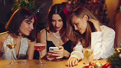 Beautiful women, friends meeting in bar, looking on mobile phone, discussing and smiling, drinking cocktails. Christmas