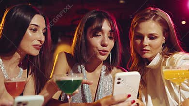 Beautiful women, friends meeting in bar, looking on mobile phone, discussing and smiling, drinking cocktails. Social