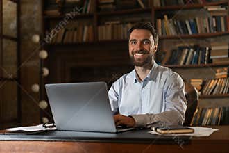 A smiling man sits at a table in the library and uses a laptop. A young scientist prepares to defend candidate or