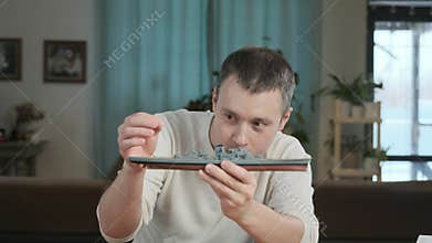 male in room at table builds a model of toy and carefully examines ship