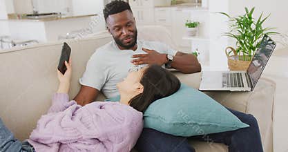 Diverse couple lying on couch and using laptop in living room