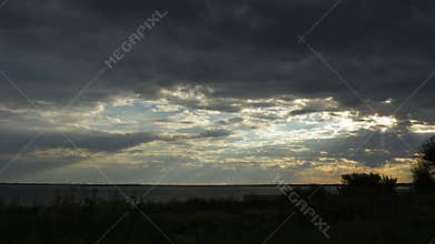 Time lapse sunset cloudscape of shore with plants shaking in wind and sunbeams breaking through grey stormy clouds