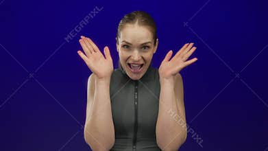 Young caucasian woman in a dark tank top covering eyes in studio in front of a deep blue backdrop; surprise astonishment amazement