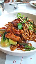 Stir-fried noodles are served in a white bowl, topped with caramelized teriyaki salmon.