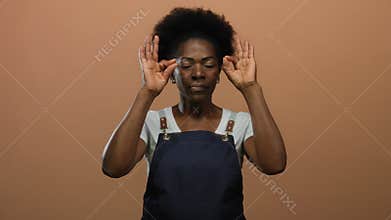 Woman wearing apron performs meditation gesture standing against isolated brown background exuding peace and concentration
