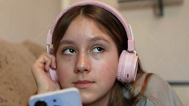 A young girl is sitting on couch and listening to music with headphones using a smartphone.