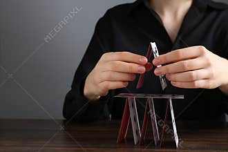 Woman building house of playing cards at wooden table against grey background, closeup. Space for text