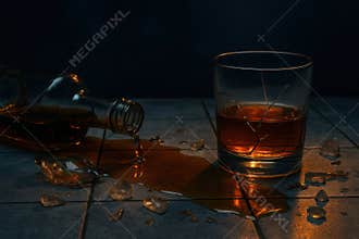 Broken glass tumbler filled with whiskey on tiled surface, spilled liquor and shattered shards illuminated in dramatic low