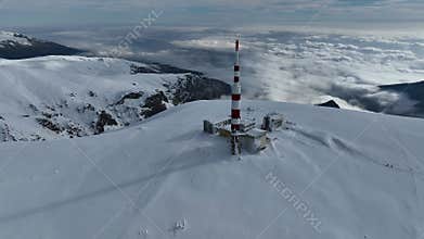 Aerial view of alpine mountain range covered in snow, with snow-capped peaks and a communications tower with antennas