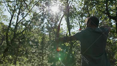 Back shot of a male archer shooting a bow and arrow in the woods on a sunny day