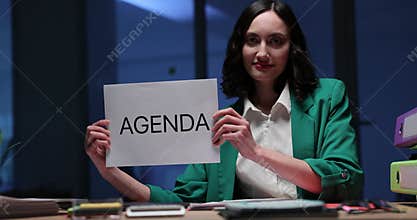 Happy businesswoman holds Agenda sign sitting at desk