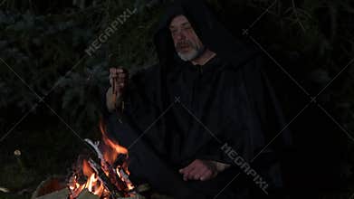 A mysterious hooded monk prays by the fire with a rosary.