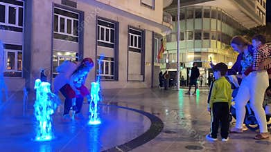Families enjoying an evening at the fountain with colorful lights in a lively city square