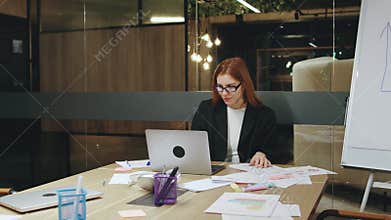 In a modern workspace, a dedicated businesswoman works late, analyzing documents on her laptop, demonstrating her