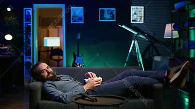 Amused man lounged on couch laughing at funny comedy movie while eating popcorn