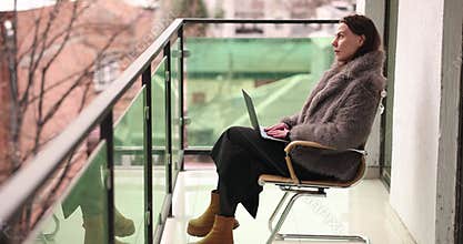 Woman author in fur jacket types on laptop sitting on balcony