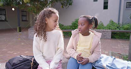 Two teenage African American girls sitting on bench with backpacks in courtyard speaking, hugging